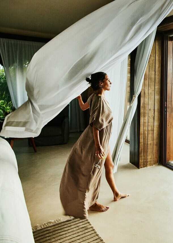 Eine Frau im langen Kleid und einem Weinglas in der Hand steht in einer Suite und blickt hinaus durch eine geöffnete Terassentür mit wehenden Gardinen.