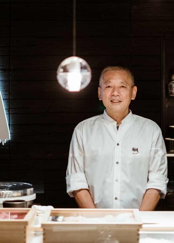 Ein Küchenchef steht hinter einer Sushibar in einem Restaurant mit einem Wandlogo Aska.