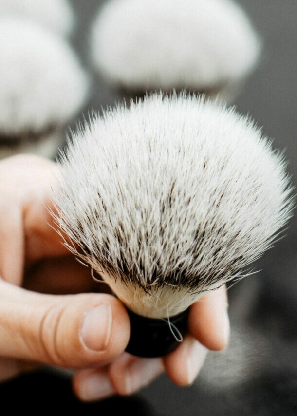 Hand hält einen weißen Rasierpinsel mit dunklen Borsten, eine zweite Hand führt eine Pinzette an die Borstenspitze.