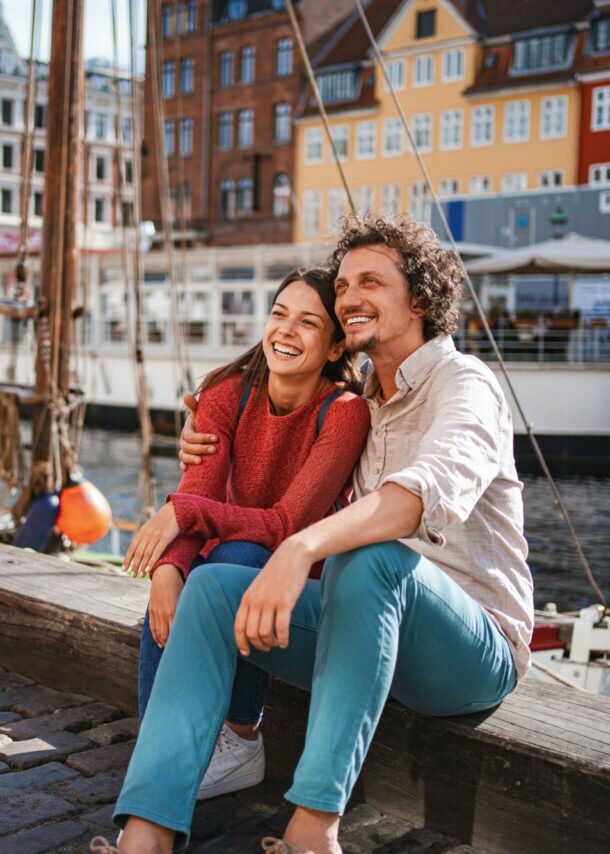 Ein lachendes Paar sitzt eng beieinander auf einer Kaimauer an einem Kanal mit bunten Häusern im Stadtzentrum bei Sonnenschein, neben ihnen stehen zwei Fahrräder auf Kopfsteinpflaster.