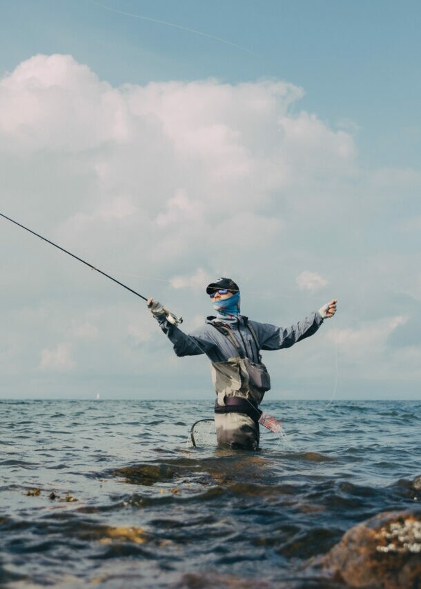 Angler steht hüfttief im Wasser zwischen Felsen und wirft eine Angelrute aus, bewölkter Himmel im Hintergrund.