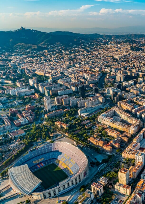 Stadtpanorama von Barcelona mit Fußballstadion aus der Luftperspektive, im Hintergrund eine Hügelkette und Meer.