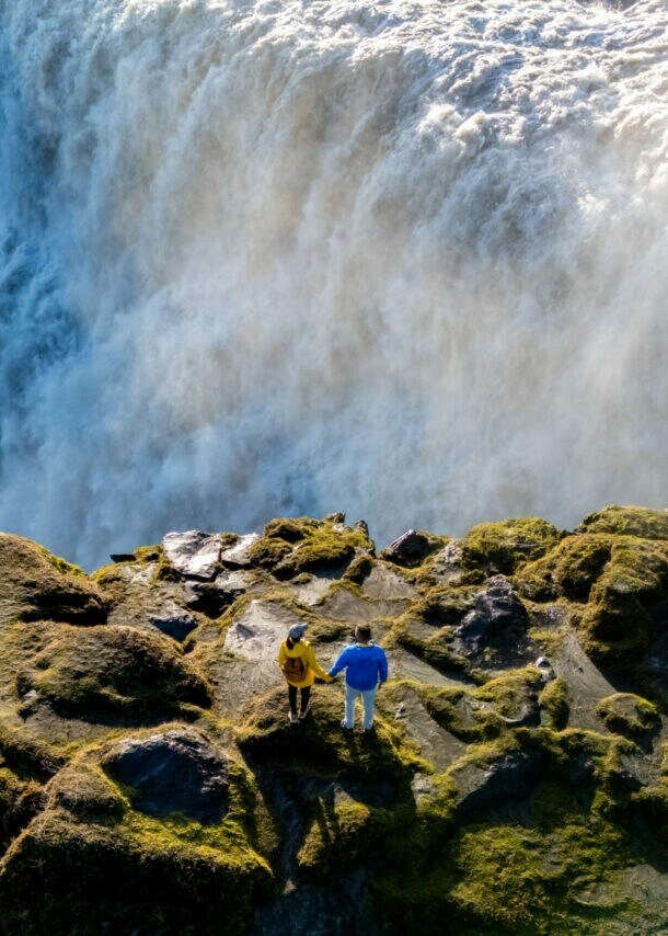 Zwei Personen stehen auf moosbedecktem Felsen vor einem großen Wasserfall mit starker Gischtentwicklung.