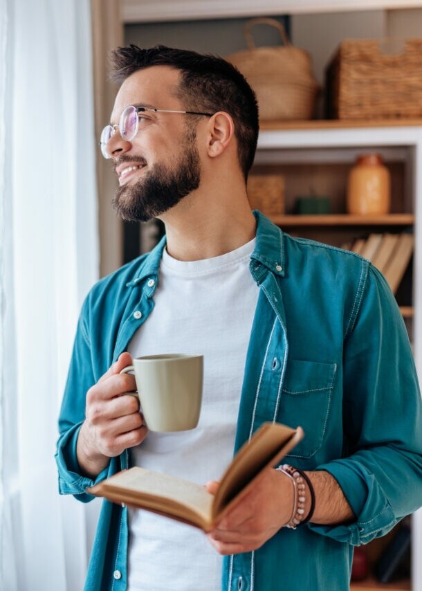 Ein Mann im Jeanshemd mit Buch und Kaffeebecher in den Händen schaut entspannt aus einem Fenster mit Vorhängen.