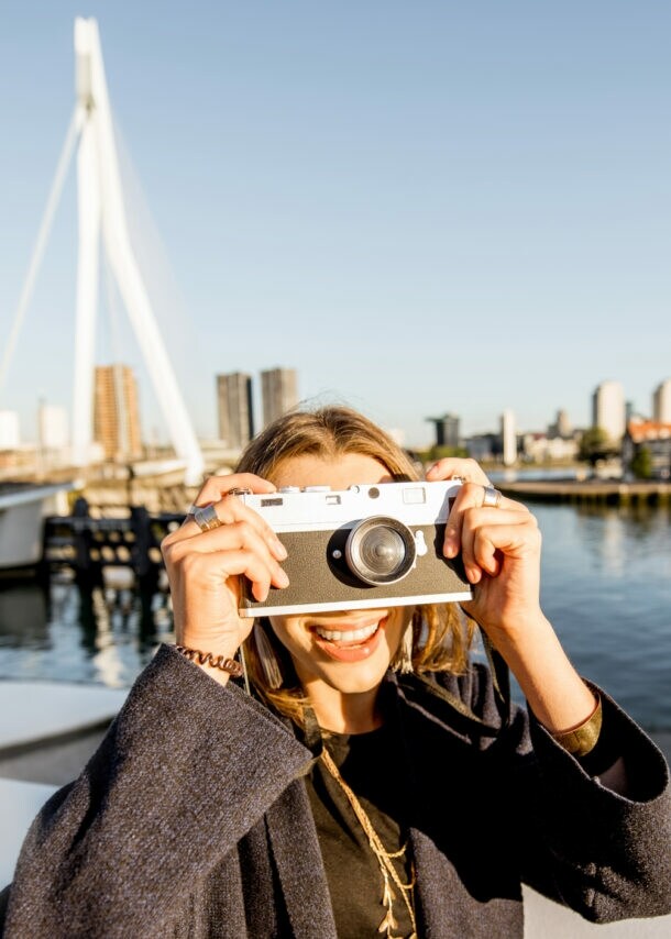 erson mit dunkelgrauem Mantel hält eine weiße Sofortbildkamera vor das Gesicht, im Hintergrund die Skyline von Rotterdam und eine Brücke über Wasser.