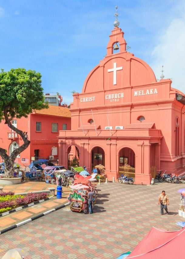 Belebter Platz mit Parkanlage, umgeben von pinkfarbenen Kolonialbauten samt Kirche unter blauem Himmel.