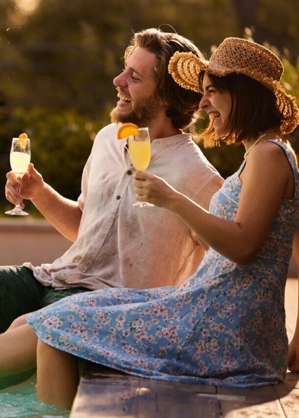 Eine Frau und ein Mann sitzen am Rand eines Pools mit einem Getränk in der Hand und spritzen mit ihren Füßen.