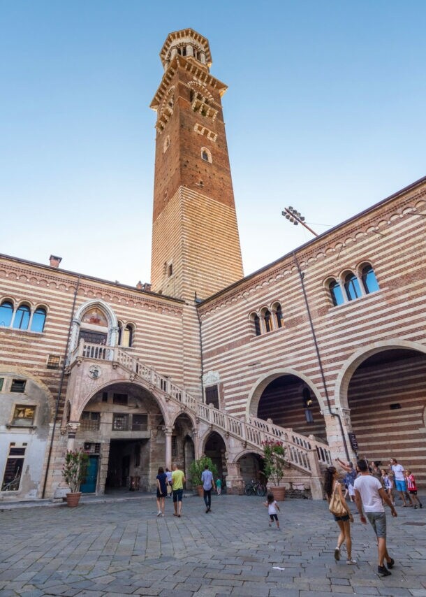Personen in einem italienischen Palastinnenhof mit gestreifter Fassade und Außentreppe unter einem Turm vor blauem Himmel.