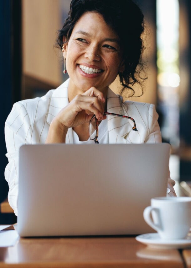 Eine Frau sitzt zufrieden lächelnd an einem Laptop an einem Tisch mit Kaffeetasse in einem Café.