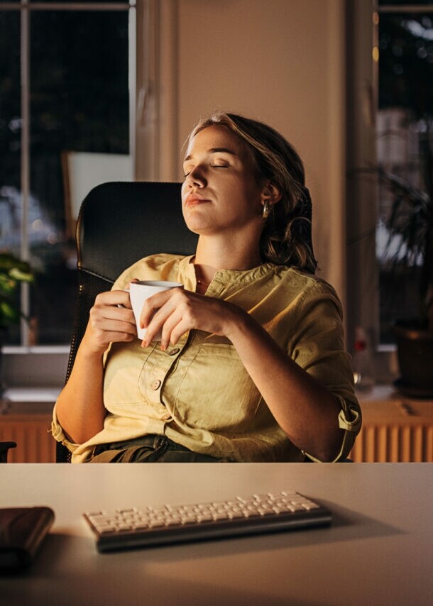 Eine Frau mit einer Tasse in den Händen lehnt mit geschlossenen Augen in einem Schreibtischstuhl an einem Schreibtisch am Abend.