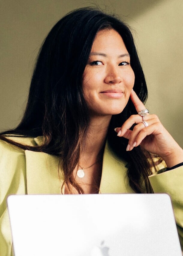 Eine Frau in einem hellgelben Blazer mit aufgestütztem Arm und einem aufgeklapptem Laptop in einem Sessel lächelt dezent in die Kamera.