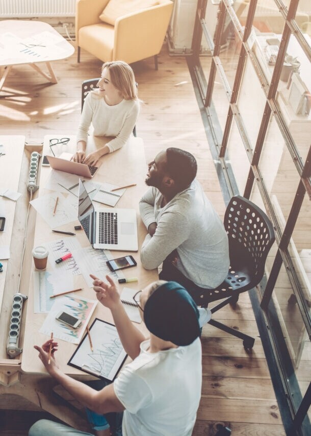 Vogelperspektive in einem Büro mit großem Tisch, an dem mehrere Personen mit Laptop sitzen und miteinander sprechen.