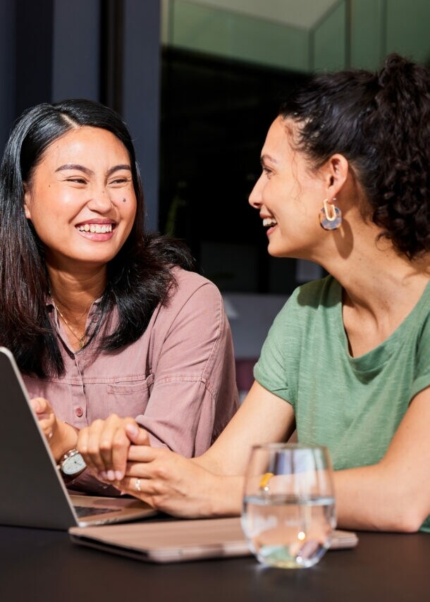 Zwei sich zugewandte lachende Frauen sitzen nebeneinander an einem Tisch, vor ihnen steht ein Laptop.