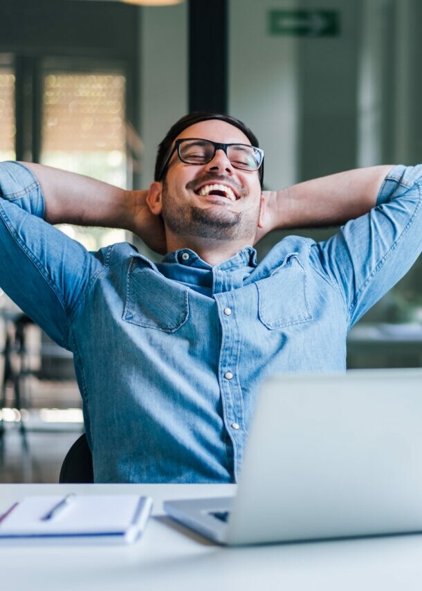 Ein lachender Mann lehnt entspannt zurück mit verschränkten Armen im Nacken auf einem Stuhl an einem Schreibtisch.
