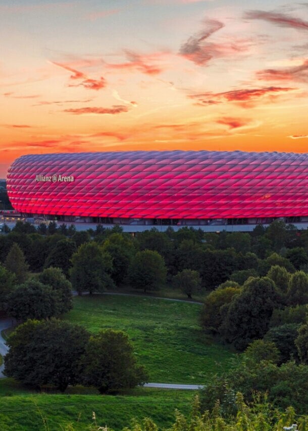 Panoramaaufnahme eines pink leuchtenden Fußballstadions unter orange gefärbtem Himmel bei Sonnenuntergang, im Vordergrund Wiesenfläche mit Wald und Kapelle.