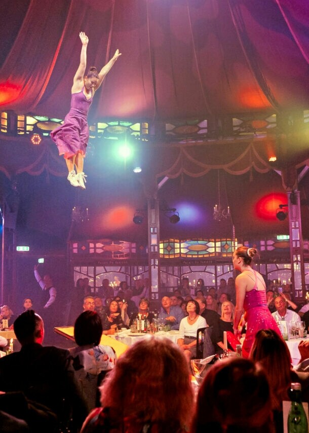 Personen sitzen beim Dinner an Tischen in einem Spiegelzelt während einer Show mit Akrobatinnen.