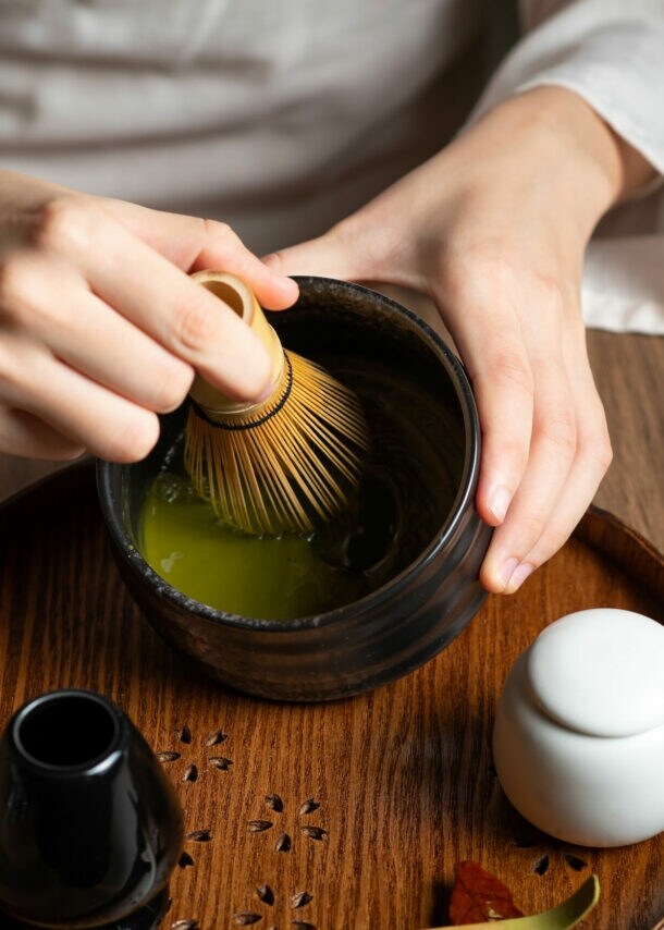 Nahaufnahme von zwei Händen bei der Zubereitung eines grünen Tees mit einem Bambusbesen in einer dunklen Keramikschale auf einem Holztisch.