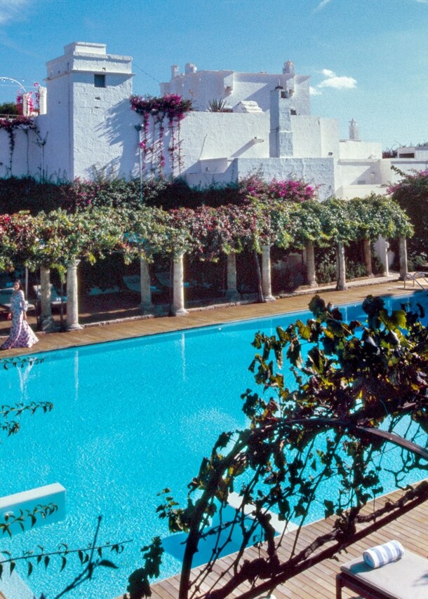 Idyllische, mit Weinranken begrünte Poolanlage in einem Hotelresort mit weißen Häusern unter blauem Himmel.