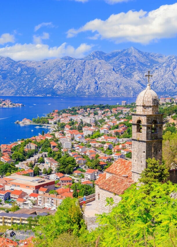 Blick auf die Stadt Kotor mit Häusern und Kirchturm, im Hintergrund Berge und eine Bucht.