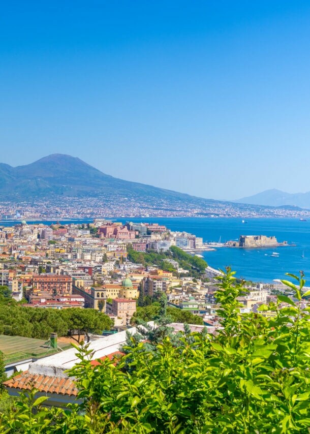 Blick von oben auf die Häuser von Neapel, im Hintergrund Meer und ein Vulkan.