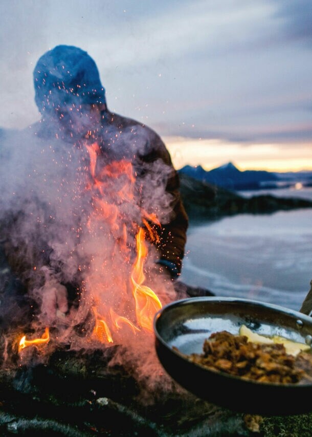 Zwei Personen bereiten Speisen in Pfannen über einem Feuer an einer schroffen Felsküste zu bei Abenddämmerung.