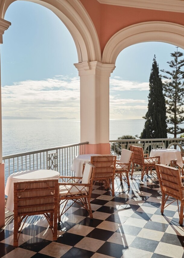 Runde Tische mit Korbstühlen auf einer Terrasse mit Meerblick durch Säulenbögen.