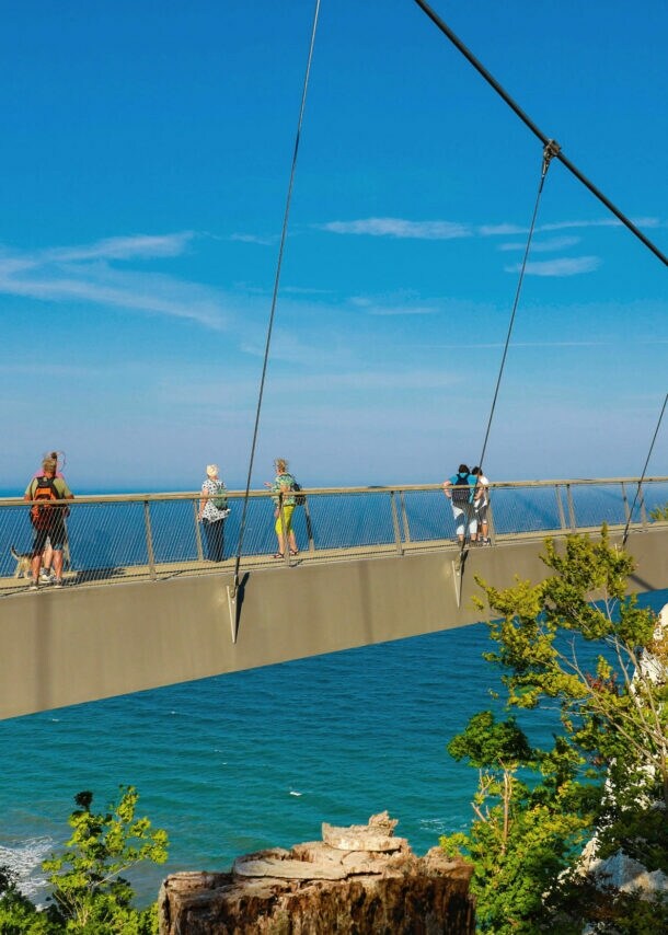 Mehrere Personen laufen auf einer schmalen Brücke zwischen Kreidefelsen am Meer unter blauem Himmel bei Sonnenschein.