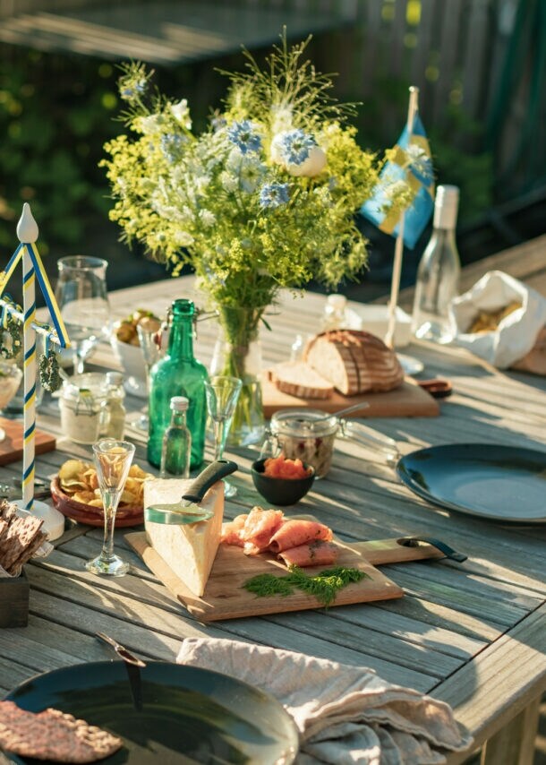 Gedeckter Holztisch für ein Mittsommer-Essen im Freien, mit Brot, gebeiztem Lachs, Käse, Eingelegtem, Getränken und frischen Blumensträußen.