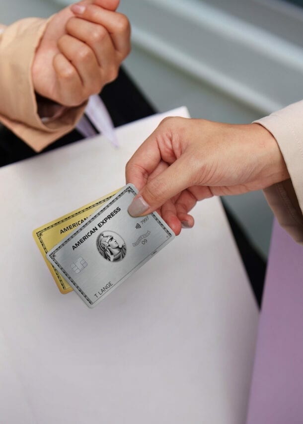 Nahaufnahme von zwei Armen mit Einkaufstüten, eine Hand hält eine goldene und eine silberne Kreditkarte von American Express.