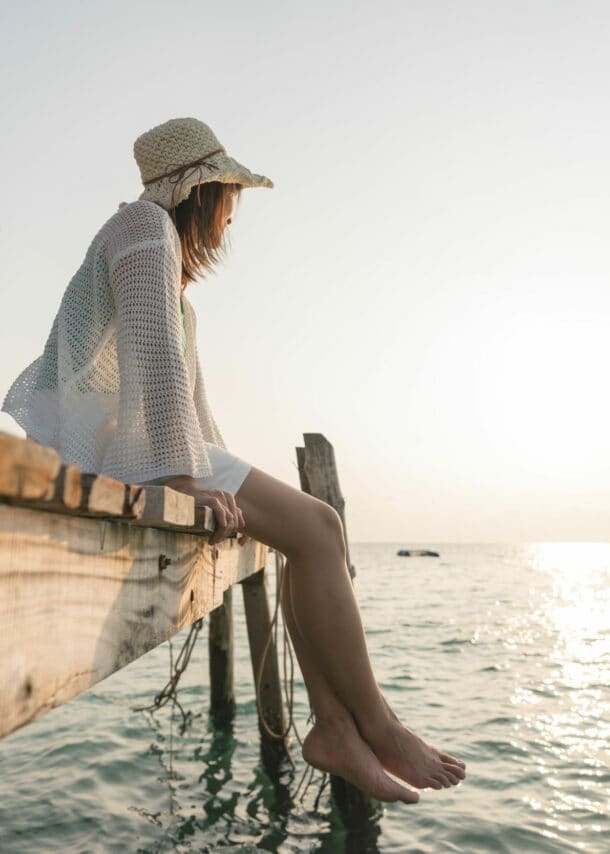 Frau mit Sonnenhut und barfuß, die auf einem Holzsteg sitzt und aufs Wasser schaut.