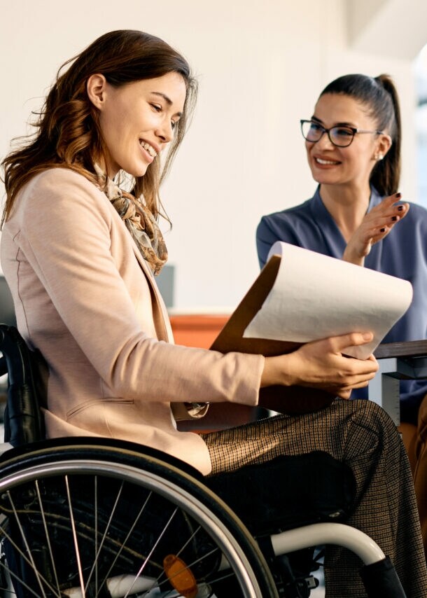 Eine Frau im Rollstuhl mit Klemmbrett in der Hand sitzt an einem Tisch neben einer weiteren Frau in einem hellen Raum.