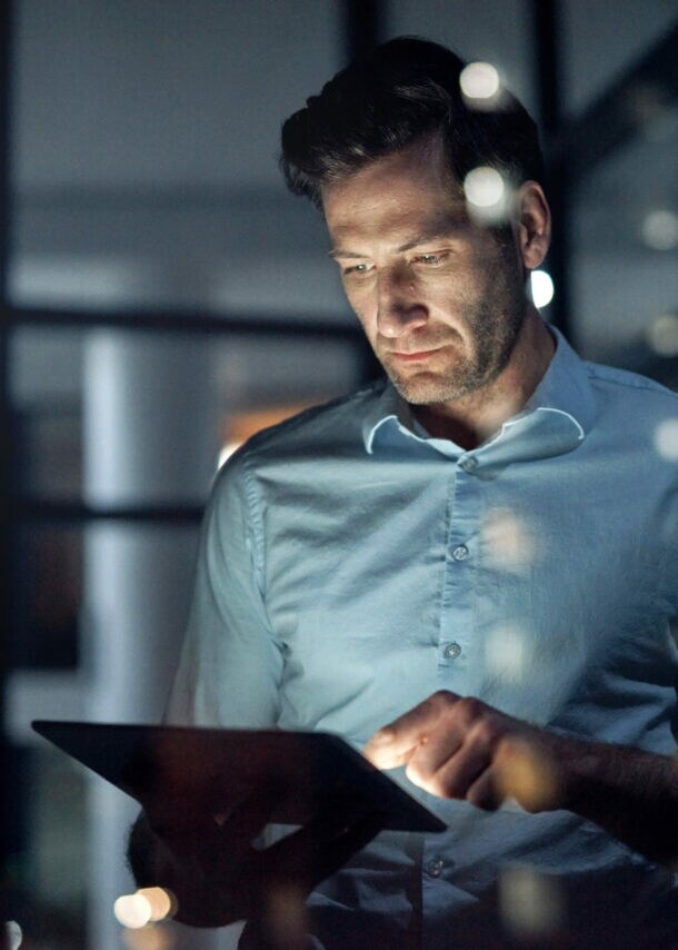 Ein Mann steht in einem dunklen Raum und hält ein Tablet in seinen Händen, dessen Displaylicht sein Gesicht anleuchtet.