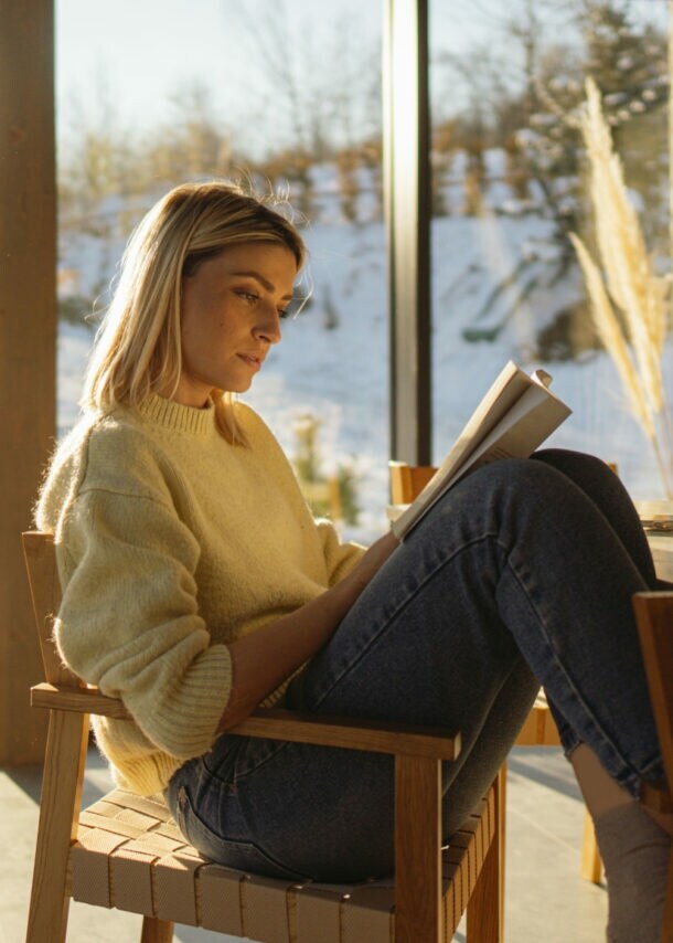 Eine Frau liest mit angewinkelten Beinen auf einem Stuhl in einem Raum mit bodentiefen Fenstern vor einer verschneiten Landschaft.
