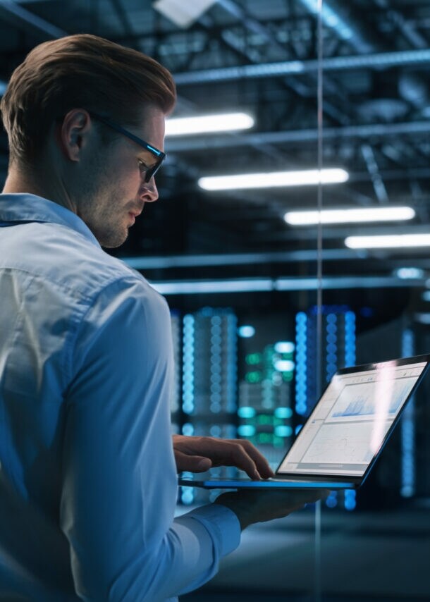 Profil eines Mannes im Hemd, der mit einem aufgeklappten Laptop in einem Rechenzentrum steht.