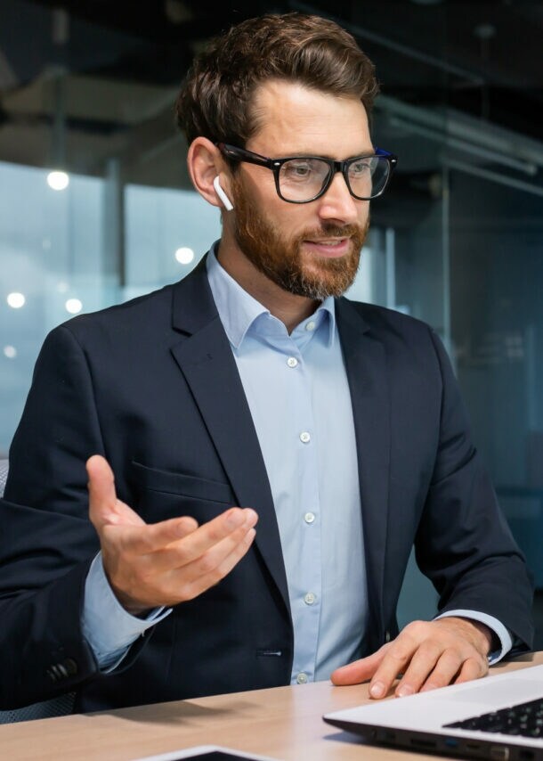 Ein Mann im Anzug und Kopfhörern in den Ohren sitzt an einem Schreibtisch vor einem aufgeklappten Laptop und gestikuliert mit der Hand in einem Raum mit Glaswand.