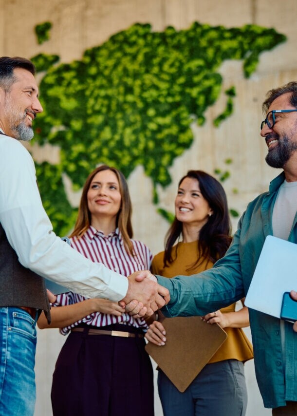 Zwei Männer mit Laptops im Arm geben sich lächelnd die Hand, daneben stehen zwei Frauen vor einer hohen Wand mit abstrakter, grüner Weltkarte.