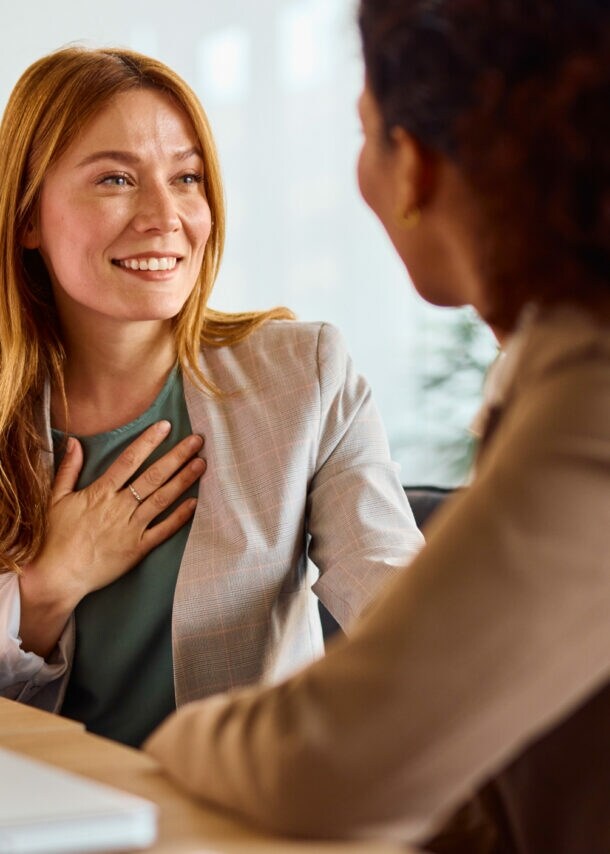 ine Frau im Blazer hat eine Hand auf die Brust gelegt und lächelt eine weitere Frau an, beide sitzen an einem Tisch, auf dem ein Tablet und ein Wasserglas stehen.