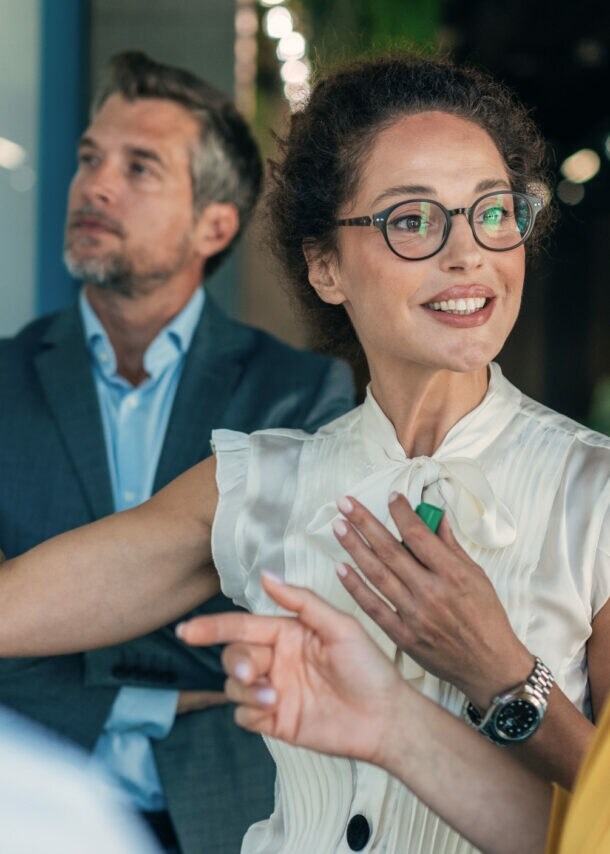 Vier Personen in Business-Kleidung stehen vor einer Wand und kommunizieren, eine Frau hält einen Stift in der Hand hoch.