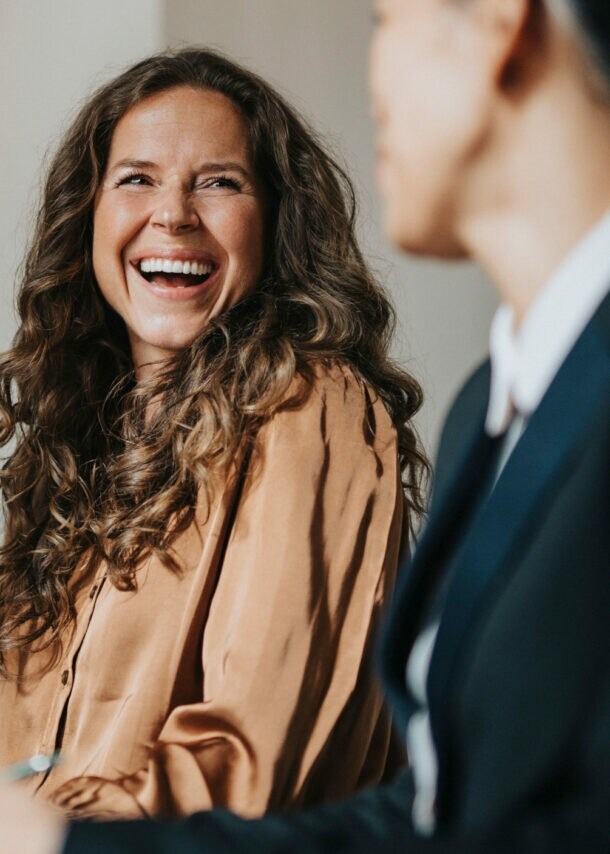 Eine lachende Frau sitzt vor einem Laptop an einem Tisch zwischen zwei weiteren Personen in Business-Kleidung während eines Meetings.