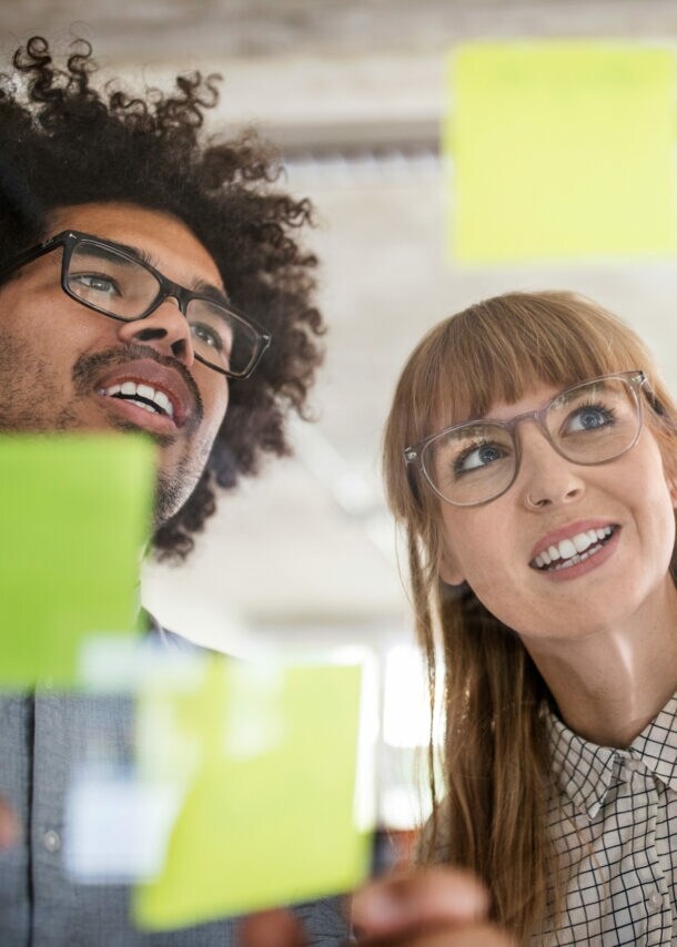 Nahaufnahme von Mann und Frau hinter einer Glasscheibe mit bunten Klebezetteln in einem Büro.