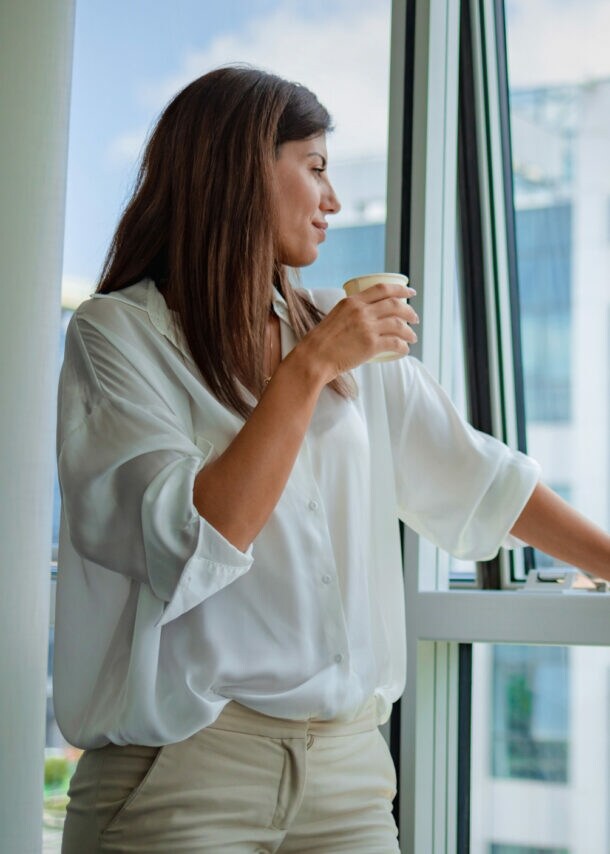 Eine Frau in weißer Bluse steht mit einem Kaffeebecher in der Hand an einem halbgeöffneten Fenster und blickt hinaus auf ein gläsernes Gebäude.