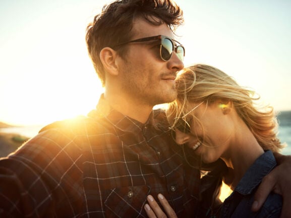 Eine Frau lehnt sich an ihren Partner, sie tragen beide eine Sonnenbrille, im Hintergrund sind das Meer und die untergehende Sonne zu erkennen