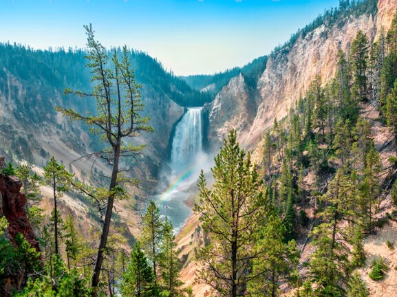Blick auf einen Wasserfall, der in die Tiefe rauscht. Davor zeichnet sich ein kleiner Regenbogen ab.