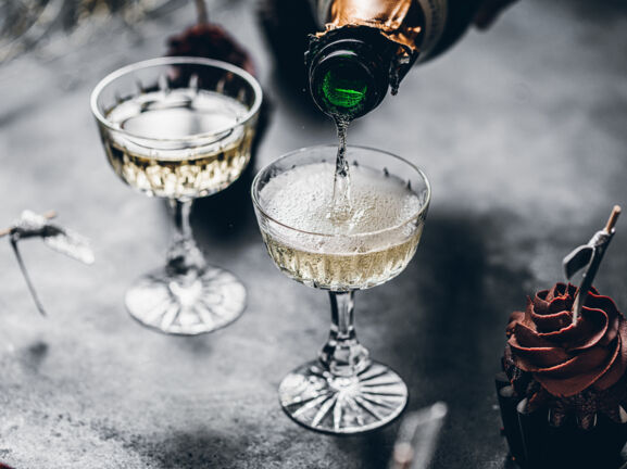 Zwei Gläser auf einem grauen Tisch werden mit Champagner gefüllt, daneben ist ein Cupcake mit Schokohaube zu sehen