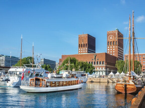 Blick auf das rote Rathaus von Oslo mit Backsteintürmen vom Wasser aus, im Vordergrund Segelboote