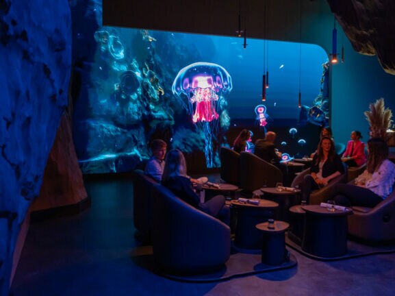 Menschen auf Sesseln in einem dunklen Raum, im Hintergrund Darstellung einer Unterwasserwelt mit leuchtenden Quallen