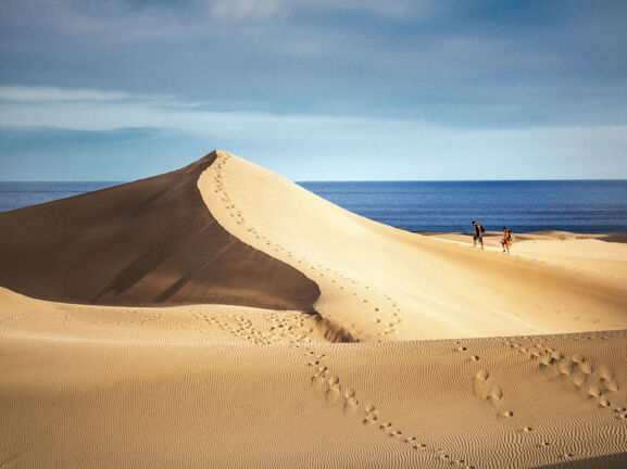 Zwei Personen wandern auf einer großen Sanddüne am Meer