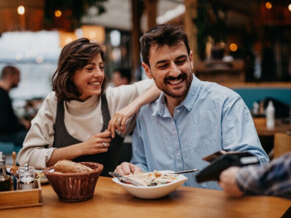 Eine Frau mit braunen Haaren und ein Mann mit Bart sitzen im Restaurant und lächeln, während er dem Kellner seine Kreditkarte zum bezahlen reicht