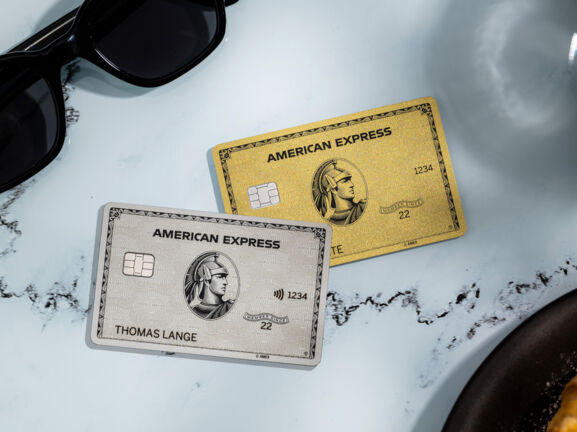 Eine silberne und eine goldene Kreditkarte von American Express liegen auf einem weißen Marmortisch neben einer schwarzen Sonnenbrille, einer Kaffeetasse und einem Teller mit Gebäck