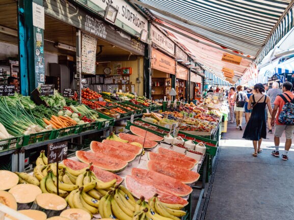 Personen auf einer Marktstraße mit Obst- und Gemüseauslagen.